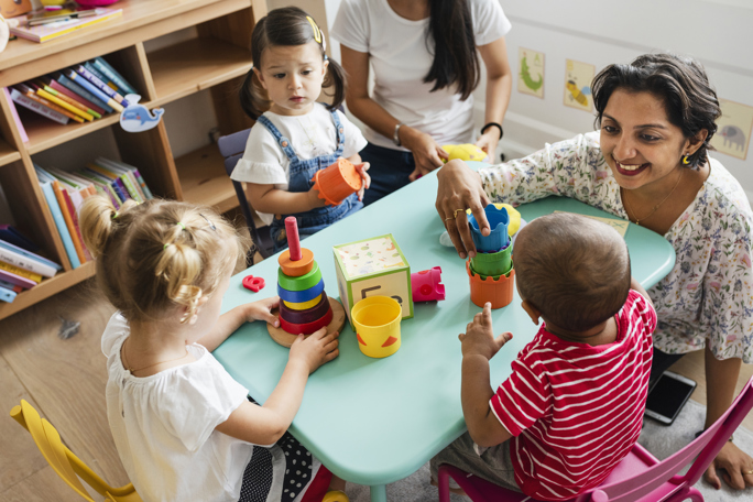 Nursery teacher engaging toddlers with stacking toys at a small activity table in a colorful classroom