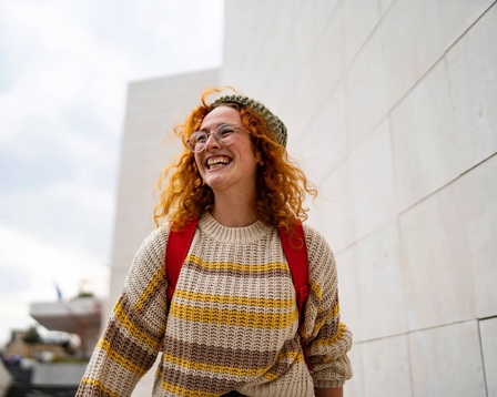A lady with bright red hair and a jumper walks along the street smiling