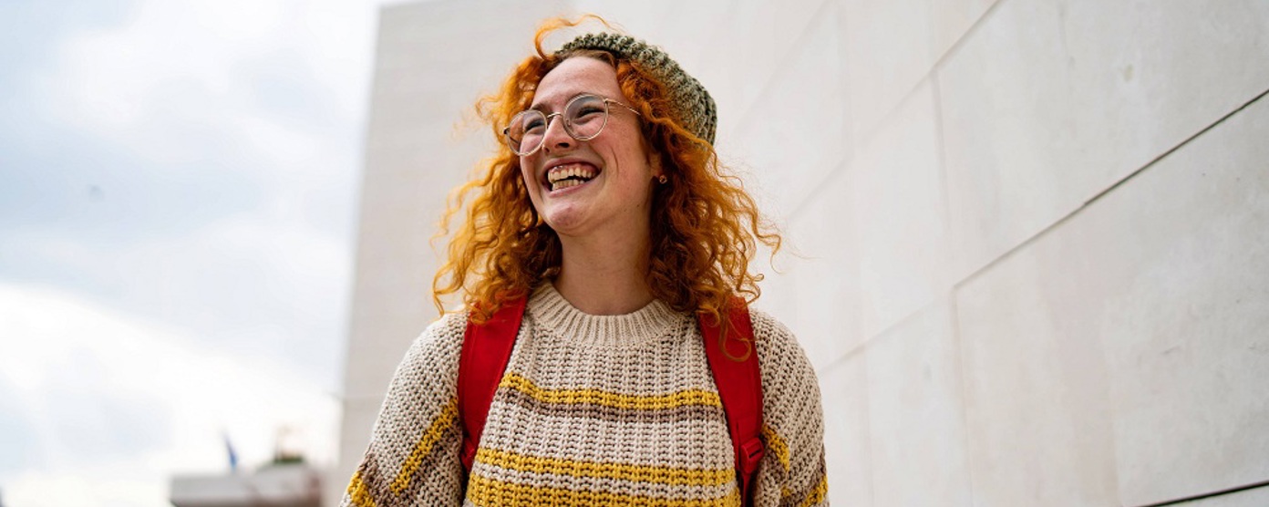 A lady with bright red hair and a jumper walks along the street smiling