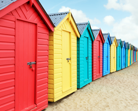 A row of brightly painted beach huts stands on sandy ground, each in different pastel and vibrant colors.