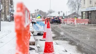 Snow-covered traffic cones and barriers line a road during heavy snowfall in an urban area.