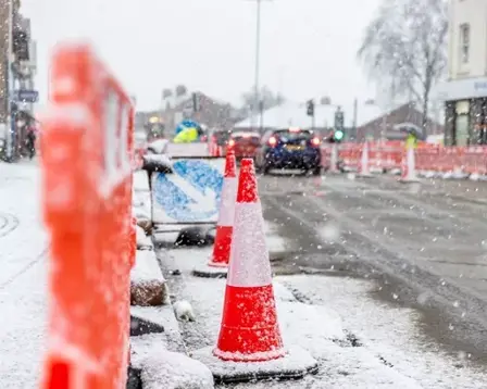 Snow-covered traffic cones and barriers line a road during heavy snowfall in an urban area.
