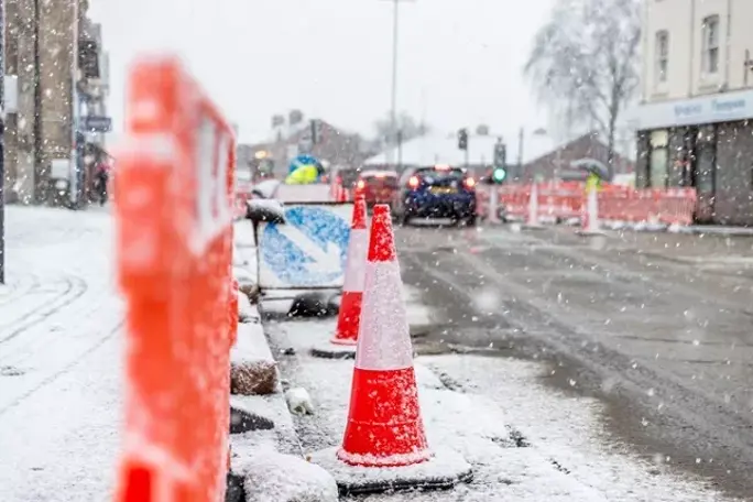 Snow-covered traffic cones and barriers line a road during heavy snowfall in an urban area.