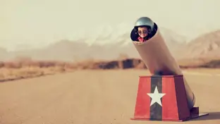 Child in a toy cannon on an empty road, wearing a helmet and sunglasses.