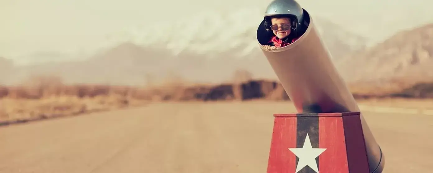 Child in a toy cannon on an empty road, wearing a helmet and sunglasses.