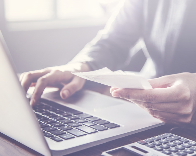 Close-up of a person managing finances at a desk, holding receipts while typing on a laptop with a calculator beside them.