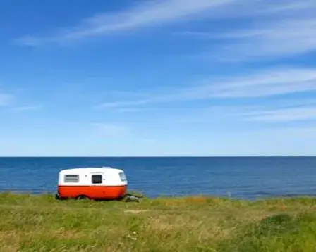 Small vintage caravan on a grassy clifftop overlooking a calm blue sea beneath a clear, streaked sky