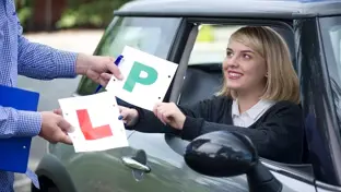 A smiling young woman in the driver's seat receives her "P" and "L" plates from an instructor, celebrating her driving milestone.