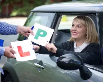 A smiling young woman in the driver's seat receives her "P" and "L" plates from an instructor, celebrating her driving milestone.