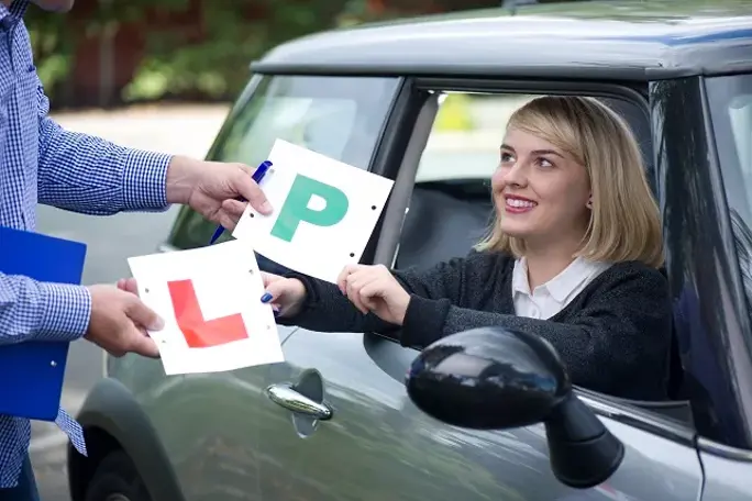A smiling young woman in the driver's seat receives her "P" and "L" plates from an instructor, celebrating her driving milestone.