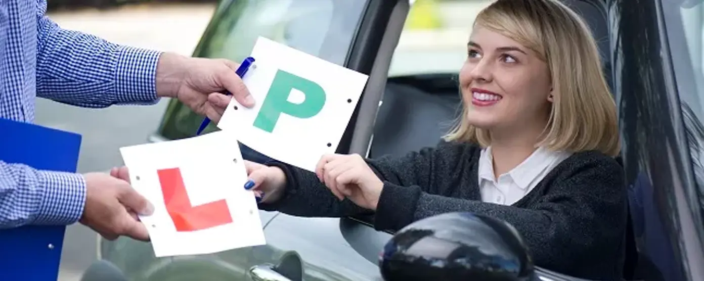 A smiling young woman in the driver's seat receives her "P" and "L" plates from an instructor, celebrating her driving milestone.