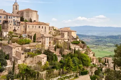 Hilltop village in France with stone buildings and cypress trees overlooking a wide valley and distant mountains.