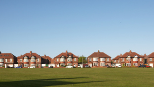 A wide row of suburban brick houses with red-tiled roofs lines the background of the image, facing a large open grassy field in the foreground.