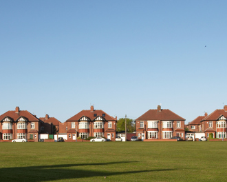 A wide row of suburban brick houses with red-tiled roofs lines the background of the image, facing a large open grassy field in the foreground.