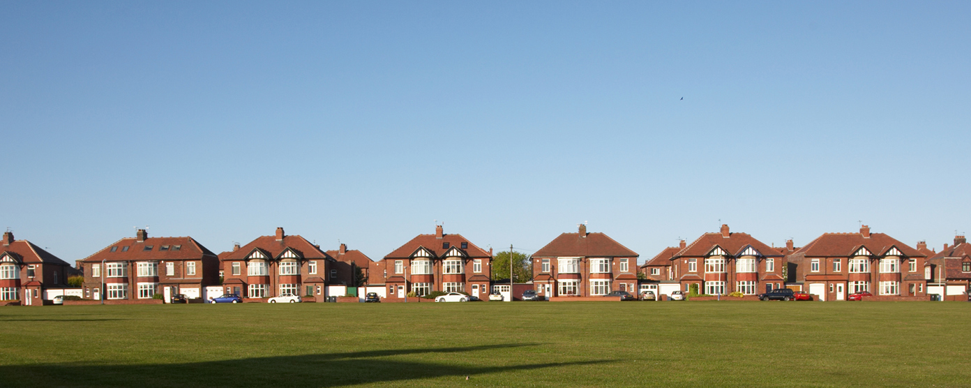 A wide row of suburban brick houses with red-tiled roofs lines the background of the image, facing a large open grassy field in the foreground.