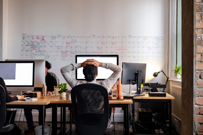 View from behind of a woman sitting at an office desk with her hands on her head