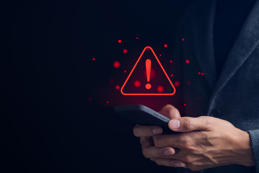 A person is holding a smartphone, with red warning icon, symbolizing a digital alert or security threat on a dark background.