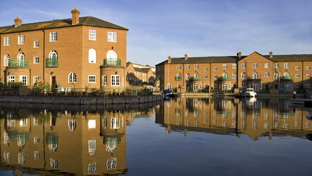 Waterside brick townhouses with green balconies reflected in a calm canal under a clear blue sky