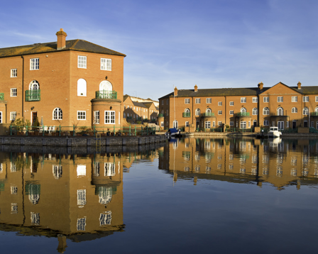 Waterside brick townhouses with green balconies reflected in a calm canal under a clear blue sky