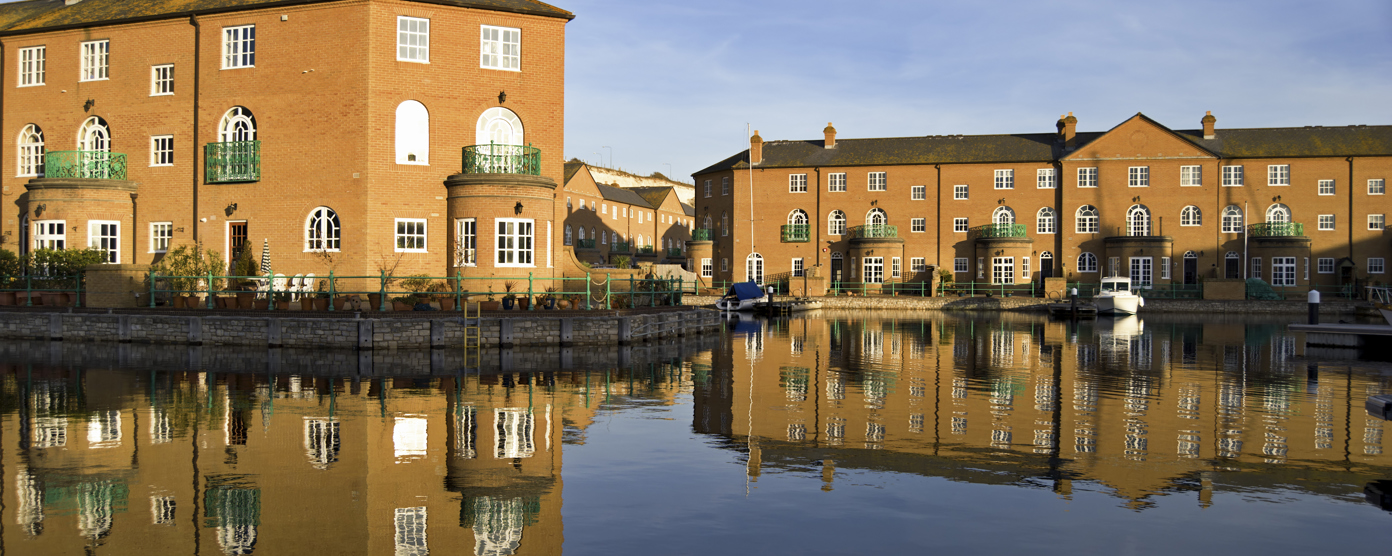 Waterside brick townhouses with green balconies reflected in a calm canal under a clear blue sky