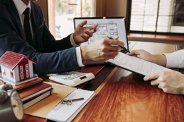 An insurance professional discusses a contract with a client, featuring a miniature house and keys on the table, related to real estate.