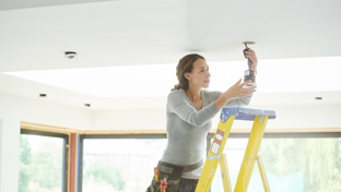 A female electrician on a ladder, focused on installing a ceiling light fixture, showcasing women's contributions in trades