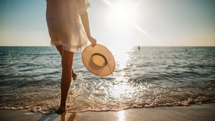 Person holding a sunhat, walking by the ocean at sunset.