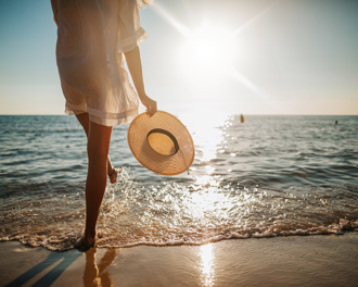 Person holding a sunhat, walking by the ocean at sunset.