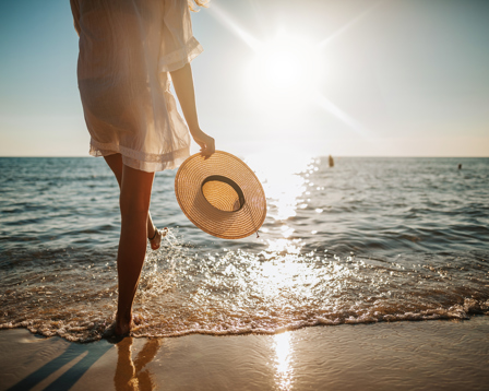 Person holding a sunhat, walking by the ocean at sunset.