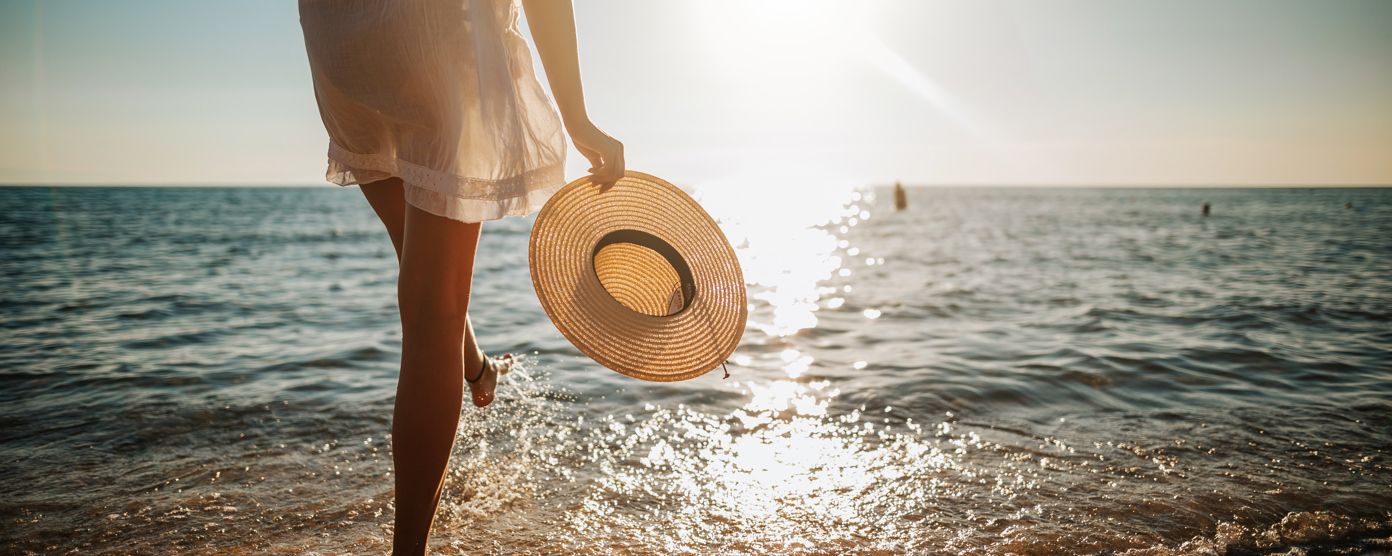 Person holding a sunhat, walking by the ocean at sunset.