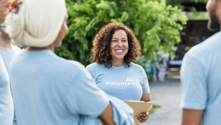 Smiling volunteer coordinator holding a clipboard and talking with a small team in light blue volunteer shirts during an outdoor meetup