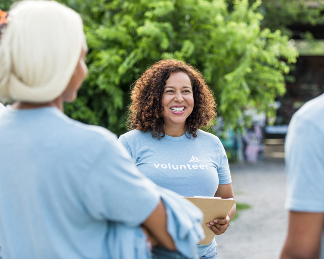 Smiling volunteer coordinator holding a clipboard and talking with a small team in light blue volunteer shirts during an outdoor meetup