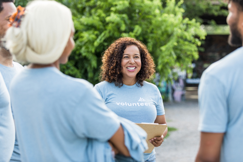 Smiling volunteer coordinator holding a clipboard and talking with a small team in light blue volunteer shirts during an outdoor meetup
