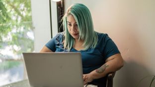 Lady with green hair and tattoos working on her laptop at a table